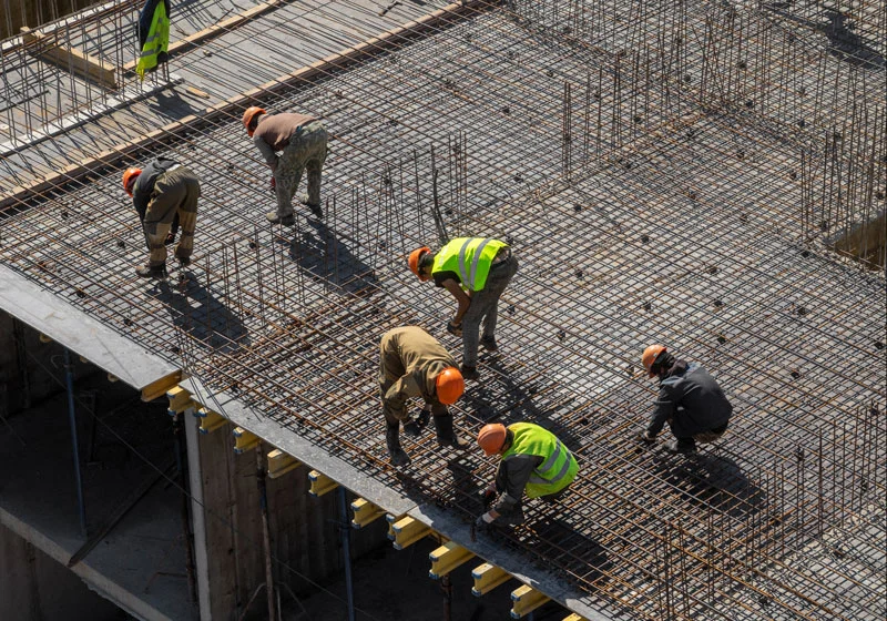 Construction workers performing tasks on an active job site with heavy equipment and building materials nearby.