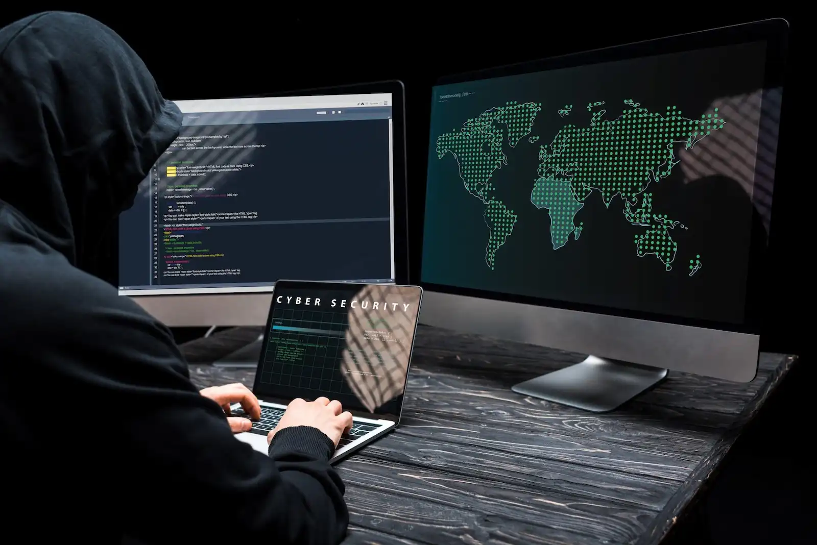 Hacker using a laptop with cyber security text on screen in a dark setup with multiple monitors